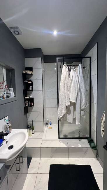 Esta é uma casa de banho moderna com azulejos cinza e brancos, um box de vidro, toalhas penduradas, um tapete preto, produtos de higiene pessoal, prateleiras e uma pia branca.