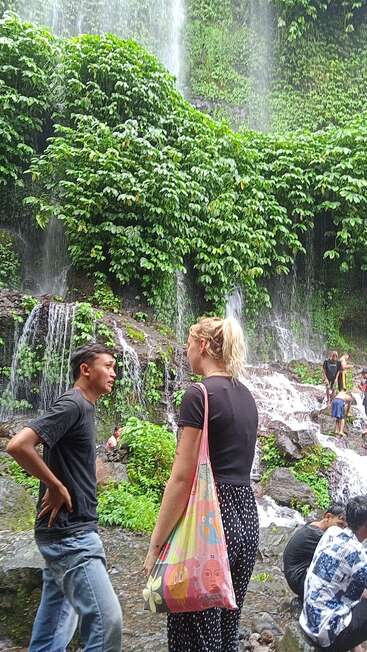 Duas pessoas estão perto de uma cachoeira verdejante, conversando. Outros apreciam o cenário natural, com água em cascata, pedras e vegetação fresca criando uma atmosfera vibrante e tranquila.