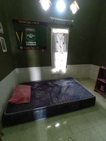 Um pequeno quarto com paredes verdes, um colchão fino no chão, um travesseiro rosa, uma janela com cortina transparente, faixas e uma pequena prateleira de madeira.