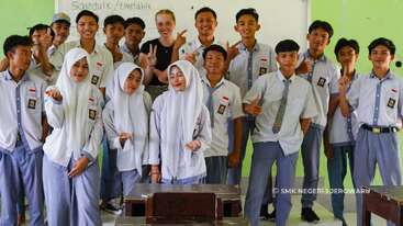 Um grupo de estudantes do ensino médio e um professor, sorrindo e posando juntos em uma sala de aula. Alguns alunos usam hijabs, todos parecem alegres e amigáveis.