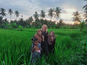 Quatro pessoas posam felizes em um campo de arroz verdejante ao pôr do sol, cercadas por palmeiras altas e natureza vibrante, criando uma atmosfera pacífica e alegre.