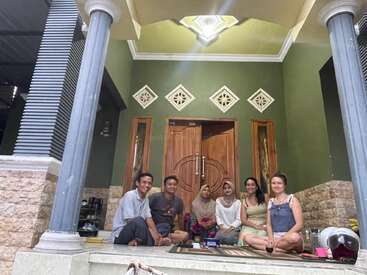 Seis pessoas estão sentadas e sorrindo juntas na varanda de uma casa com paredes verdes, pilares decorados e uma porta de madeira, parecendo relaxadas e felizes.