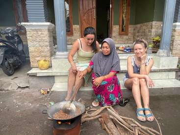 Três mulheres sentadas juntas do lado de fora de uma casa, cozinhando em uma fogueira com lenha, sorrindo enquanto preparam a comida, desfrutando da companhia umas das outras em um ambiente descontraído.