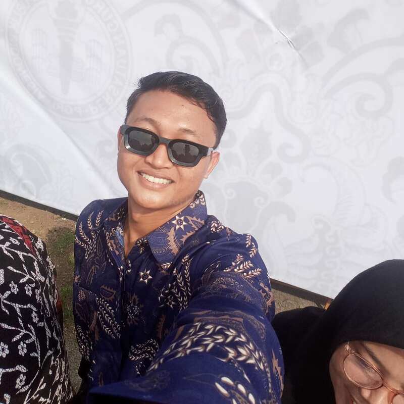 Um homem sorridente, usando óculos escuros e camisa tradicional de batique, tira uma selfie ao ar livre. Duas outras pessoas estão parcialmente visíveis. O plano de fundo é um banner branco estampado.