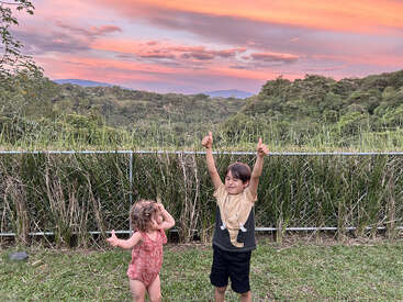 Deux enfants joyeux se tiennent debout sur l'herbe, levant le pouce. Derrière eux, des arbres et des buissons verdoyants. Le ciel resplendit de magnifiques teintes orange et violettes au coucher du soleil.