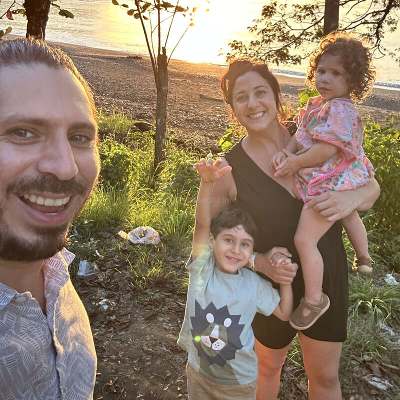 Une famille heureuse de quatre personnes pose en plein air au coucher du soleil près d'une plage. Les parents et les enfants sourient, la verdure et la lumière du soleil créant une atmosphère chaleureuse.