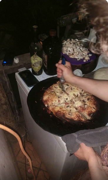 A person cuts a homemade pizza topped with mushrooms in a dimly-lit area. Nearby are bottles, chopped mushrooms, a phone, and some kitchen tools. Cozy atmosphere.
