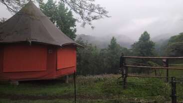 A round, orange tent stands near a rustic wooden fence, surrounded by lush greenery and misty mountains in the background beneath an overcast sky. Serene atmosphere.