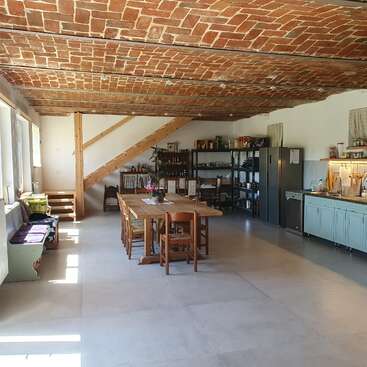 Dieses Bild zeigt eine geräumige Küche im Landhausstil mit einer Ziegeldecke, einem Esstisch aus Holz, offenen Regalen, modernen Geräten, großen Fenstern und einer Holztreppe. Gemütliche Atmosphäre.