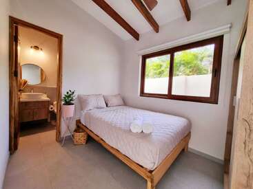 This cozy bedroom features a neatly made bed, wooden beams, potted plant, large window with outdoor view, and an ensuite bathroom with a modern sink and mirror.