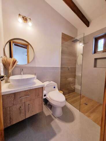 This modern bathroom features a glass-enclosed shower, vessel sink, wooden vanity, round mirror, toilet, vanity lighting, neutral tones, high ceiling, and minimalist decor.