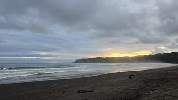 Ein ruhiger Strand bei Sonnenuntergang mit sanften Wellen, die auf den dunklen Sand rollen. Der Himmel ist bewölkt, doch in der Nähe eines entfernten, bewaldeten Hügels bricht goldenes Sonnenlicht durch.