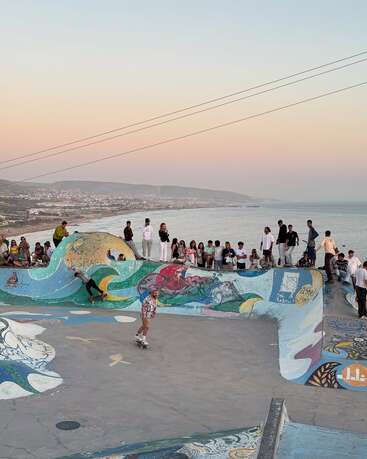 Ein farbenfroher Skatepark am Meer voller Menschen, von denen einige skaten und andere zuschauen. Lebendige Graffiti zieren die Rampen. Der Sonnenuntergang und das Meer bilden eine friedliche Kulisse.
