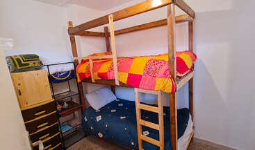 Dieses Bild zeigt ein kleines Schlafzimmer mit Etagenbetten aus Holz. Das obere Bett hat eine bunte Steppdecke und das untere eine blaue Decke. Es gibt Regale und Schubladen.