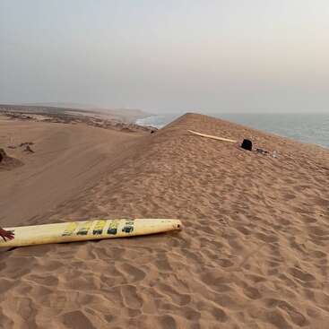 Eine Sanddüne trifft unter einem dunstigen Himmel auf den Ozean. Zwei Surfbretter, ein Rucksack und eine Wasserflasche liegen auf dem strukturierten Sand und schaffen Einsamkeit.