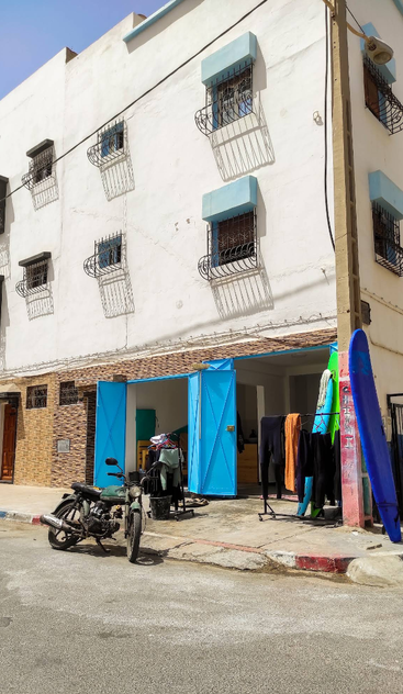 Un edificio blanco con toldos azules en las ventanas y puertas azules abiertas. En el exterior, a la luz del sol, se ve una motocicleta, una tabla de surf y trajes de neopreno secándose en un tendedero.