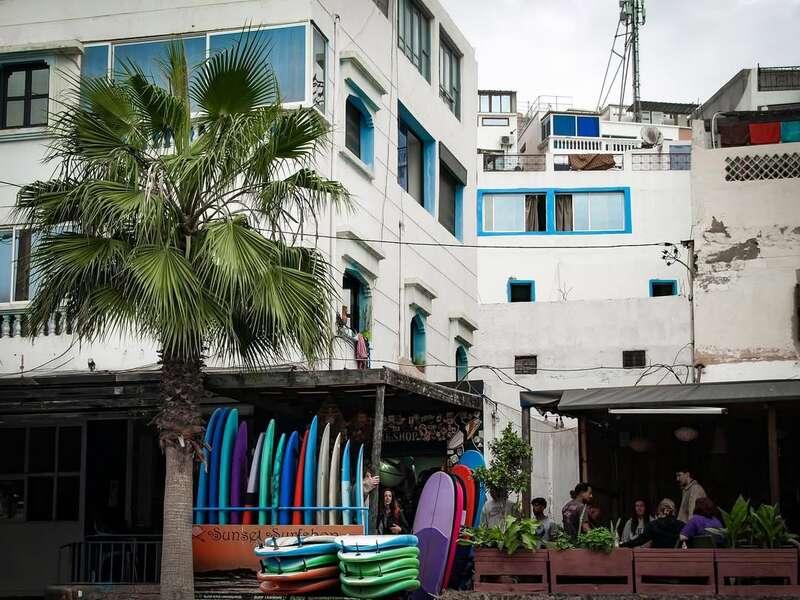 Vor einem weißen Gebäude mit blauen Fenstern stehen bunte Surfbretter neben einer Palme. Die Menschen versammeln sich in einem schattigen Außenbereich und schaffen eine entspannte Atmosphäre.