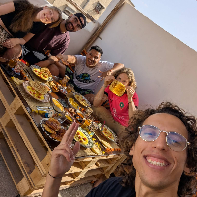 Cinco amigos disfrutan juntos de un alegre desayuno al aire libre, sentados alrededor de una rústica mesa de madera llena de platos coloridos, té y comida tradicional, sonriendo y posando felices.
