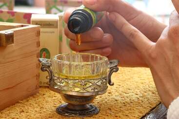 Eine Person gießt gelbes ätherisches Öl aus einer kleinen Flasche in ein verziertes Glasgefäß, das auf einer dekorativen Oberfläche neben einer Holzkiste steht.