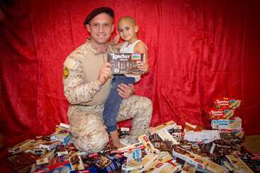 Ein lächelnder Soldat in Tarnkleidung kniet neben einem jungen, kahlen Kind. Sie halten eine Loacker-Schokoladenschachtel, umgeben von vielen ähnlichen Schachteln, vor einem roten Hintergrund.