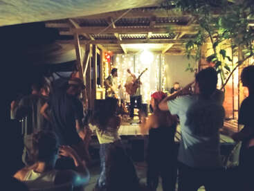 A lively, intimate outdoor concert at night, featuring a small band on a cozy, decorated stage. Audience members are dancing, enjoying music under warm, festive lights.
