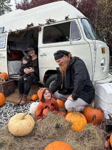 A family celebrates autumn, sitting by a white camper van surrounded by pumpkins and hay. The child wears an orange pumpkin costume, creating a festive Halloween scene.