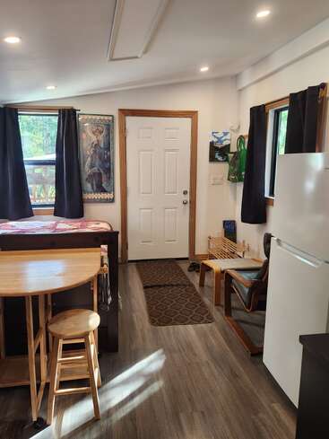 A cozy, sunlit room with wood floors features a bed, small table with stool, chair, mini fridge, wall art, dark curtains, and a front door.