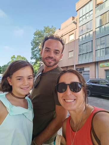 A cheerful group of three—two adults and a girl—smile outdoors on a sunny day, standing in front of modern buildings and a parked black car.