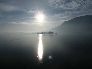 The image depicts a serene lake scene with a mountainous backdrop, featuring a bright sun shining down on the water's surface. The sky is cloudy.