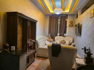 A cozy living room with cream sofas, ornate ceiling lights, dark curtains, wooden furniture, a globe, TV, and decorative items, creating a warm, inviting atmosphere.