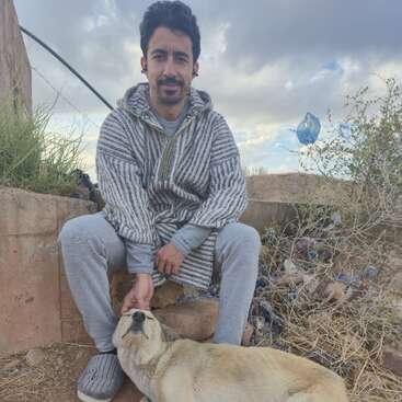 A man in a striped hoodie sits outdoors on a stone ledge, gently petting a relaxed dog. The sky is cloudy and plants surround them.