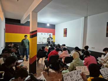 Une salle de classe avec de nombreux étudiants assis face à un instructeur à l'avant. Les murs sont décorés aux couleurs du drapeau allemand. Les étudiants tiennent des documents imprimés.