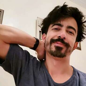 A man with messy hair and a beard smiles gently, wearing a casual t-shirt. He poses indoors, hand behind his neck, and appears relaxed and comfortable.