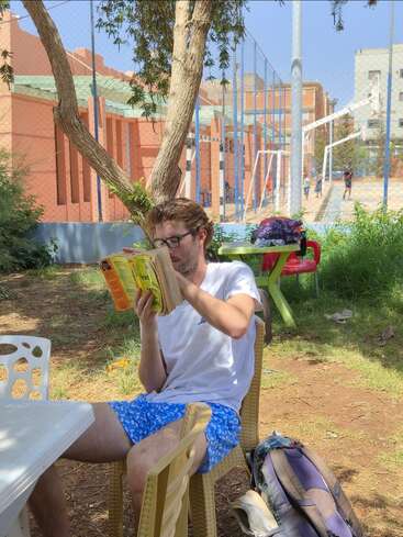 Un homme portant des lunettes est assis à l'extérieur, sous un arbre, en train de lire un livre. Il porte une chemise blanche et un short bleu, et est assis à côté d'une table et d'un sac à dos.