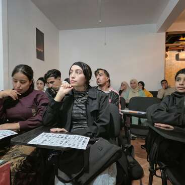 A diverse group of students sits attentively in a classroom, focused on a lesson. They have papers in front of them and appear engaged in learning.