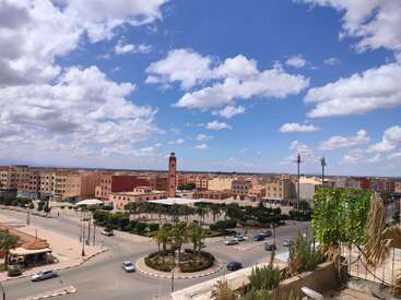 The image shows a bright cityscape with pastel-colored buildings, a central clock tower, palm trees, roundabouts, scattered cars, and a blue sky filled with clouds.