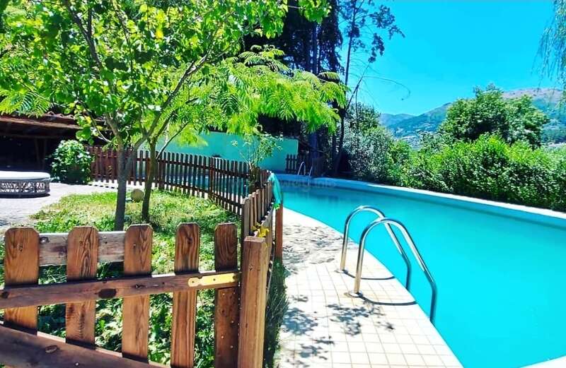 A narrow, curved swimming pool sits beside a wooden fence and lush greenery, under a bright blue sky, surrounded by trees and distant hills. Peaceful atmosphere.