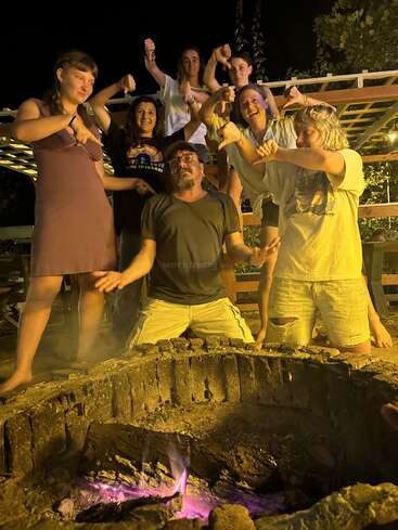 A group of people gathers around a campfire at night, playfully pointing and giving thumbs down to the man in the center, creating a fun atmosphere.