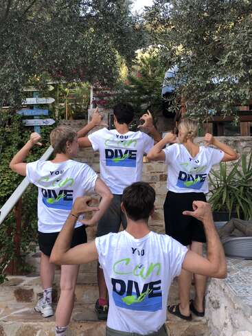 Four people stand on stone steps, backs facing the camera, wearing matching "You Can Dive" t-shirts and flexing their arms, surrounded by lush greenery and signs.