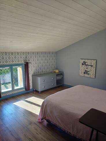 This cozy bedroom features a slanted white ceiling, patterned wallpaper, large window with curtains, wooden floors, a bed with a pink cover, and a gray dresser.