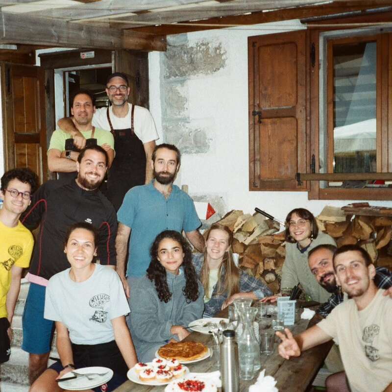 Un groupe de onze personnes se réunit autour d'une table rustique en bois, souriant, savourant la nourriture et la compagnie des autres à l'intérieur, avec du bois de chauffage et des fenêtres en bois douillettes derrière eux.