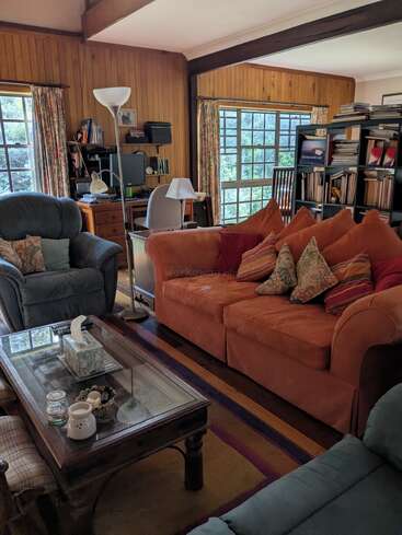 A cozy living room with wood-paneled walls, large windows, orange and blue sofas, a glass coffee table, bookshelves, a desk, and colorful cushions.