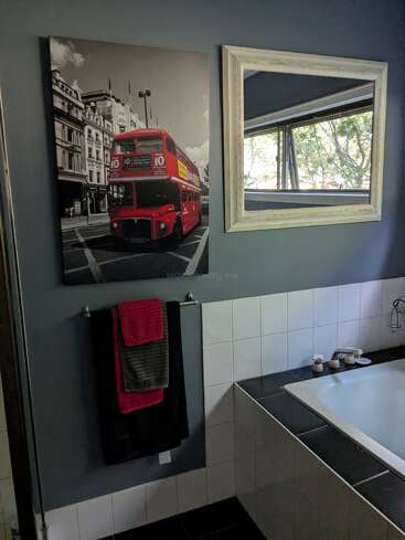 This bathroom features a bathtub, white tiles, dark walls, a mirror, a towel rack with folded towels, and wall art of a red double-decker bus.