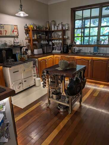 This cozy kitchen features wooden cabinets, open shelves with jars, a central island with pots and utensils, vintage stove, large window, and a rustic, homey atmosphere.