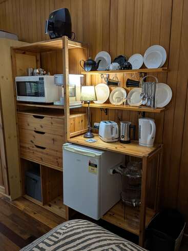 The image shows a compact wooden kitchen corner with a microwave, mini fridge, toaster, kettle, plates, mugs, utensils, an air fryer, and a lamp.