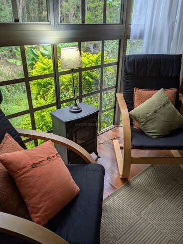 A cozy sunroom with two cushioned wooden chairs, a small black table lamp, large windows overlooking greenery, wooden flooring, and soft pillows for comfort.
