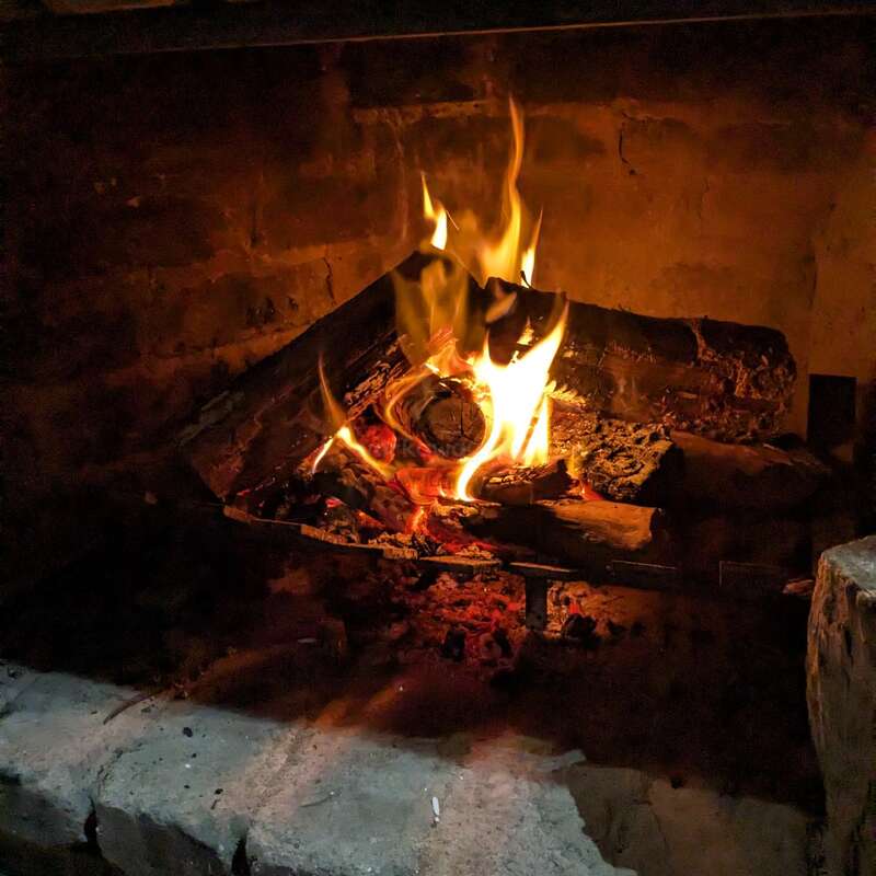 A cozy fireplace with burning logs glows warmly. Bright orange and yellow flames flicker, casting light on rustic bricks and creating a comforting, inviting atmosphere.