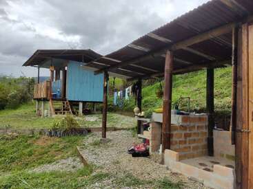 Una pequeña y rústica granja con una casa de madera azul sobre pilotes, una cocina al aire libre y ropa tendida, rodeada de vegetación, bajo un cielo nublado en plena naturaleza rural.