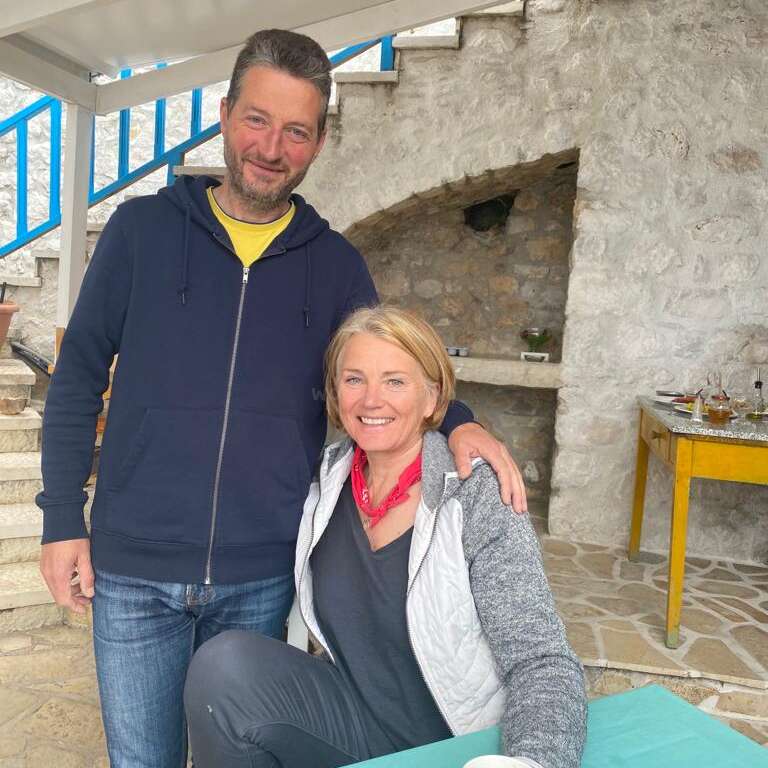 Das Bild zeigt einen Mann und eine Frau, die für ein Foto posieren, wobei der Mann hinter der Frau steht, die auf einem Steinboden sitzt.