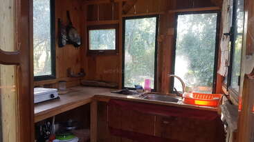 This image shows a cozy wooden kitchen with large windows, a sink, dish rack, stove, and hanging utensils. Sunlight streams in, creating a warm, inviting atmosphere.